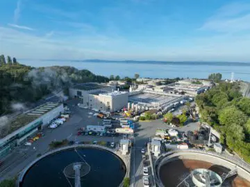Aerial view of a coastal wastewater treatment plant with circular tanks, surrounded by greenery and overlooking a serene body of water.