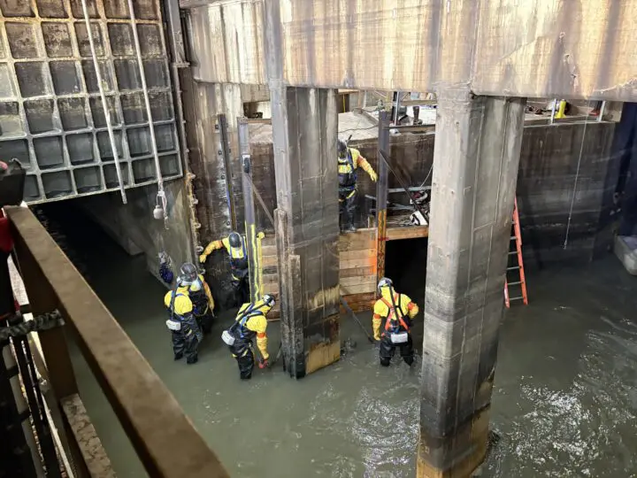 Workers in safety gear assess a water-filled structure. Industrial environment with ladders and machinery.