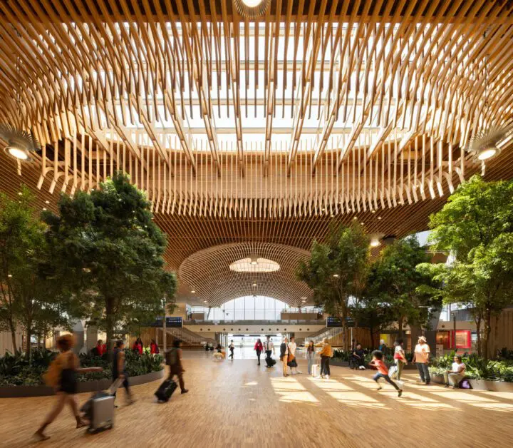 Modern atrium with unique wooden ceiling, natural light, and indoor trees. People walking and relaxing in an open, airy space.