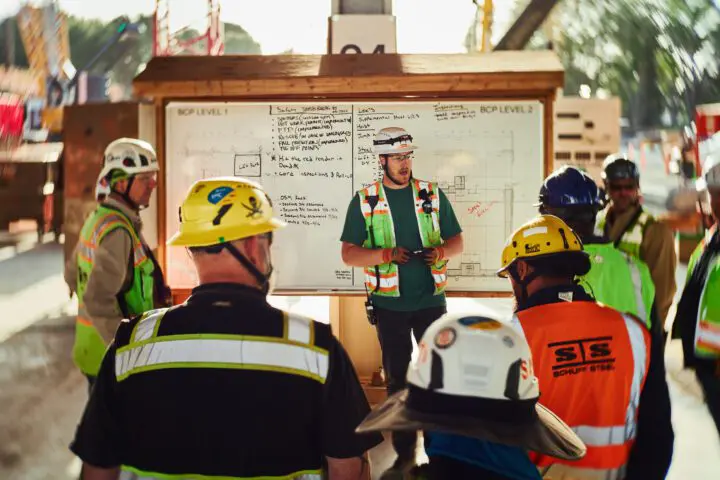 Construction team briefing at a site with individuals in safety gear and helmets, standing around a whiteboard.
