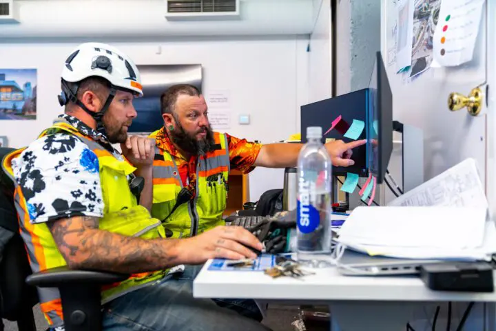 Two construction workers in safety vests and helmets collaborate at a computer, reviewing blueprints and notes in an office setting.