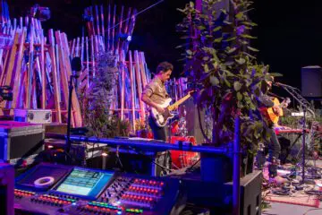 Live outdoor concert with musicians playing guitar and bass on a vibrant stage, surrounded by colorful lights and greenery.