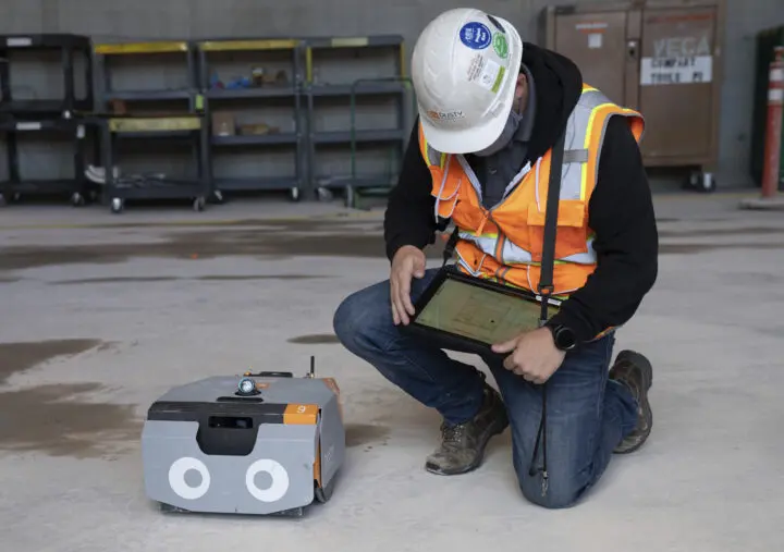 Worker using tablet to control robotic device on construction site, wearing safety gear and helmet, checking floor plans.