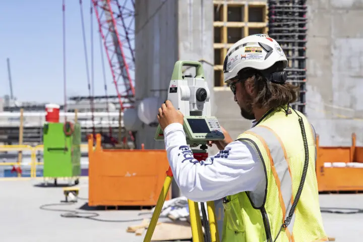 Surveyor using a total station on a construction site; wearing a hard hat and safety vest. Equipment in the background.