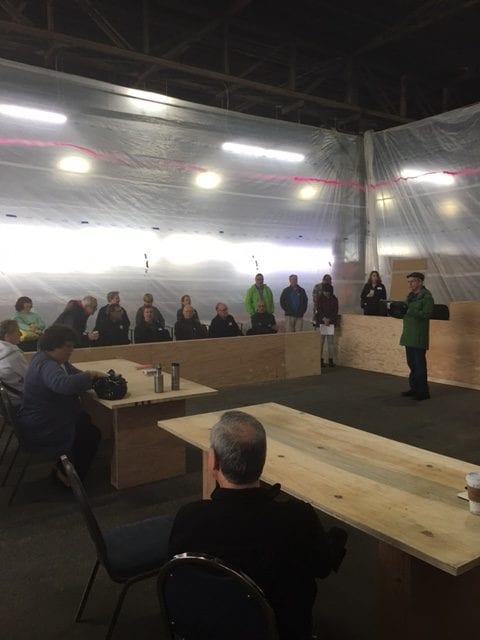 Group meeting in a large industrial space with a speaker addressing individuals seated at tables and chairs.